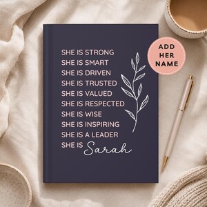 May include: A navy blue journal with the words "She is Strong" and other empowering phrases in white. A floral design and the name "Sarah" are also featured. A pen and a cup of coffee are visible.