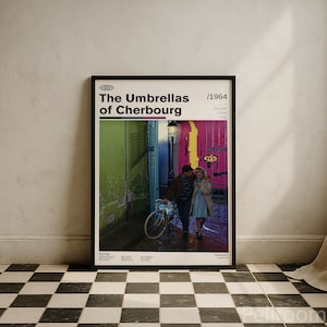 May include: A framed film poster for The Umbrellas of Cherbourg from 1964. The poster features a central image of two people standing near a bicycle on a street with bright green and pink walls. The text includes the title, year, and credits.