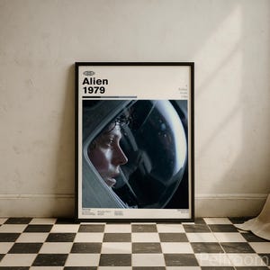 May include: Framed movie poster for the film "Alien" from 1979. The poster shows a close-up of a person in a spacesuit helmet. The title "Alien 1979" is printed at the top in a bold font. The poster is in a black frame.