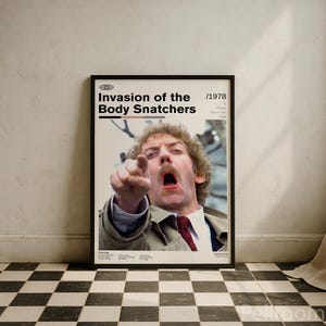 May include: Framed movie poster for "Invasion of the Body Snatchers" (1978). The poster features a man pointing with an open mouth. The title is in bold white letters, with the year in the upper right corner. The poster is in a black frame.