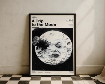Puede incluir: Póster enmarcado en negro con el título de la película "A Trip to the Moon" con estética vintage. El póster muestra una imagen en blanco y negro de la luna con una cara, observada a través de un telescopio. El póster se exhibe contra una pared blanca.
