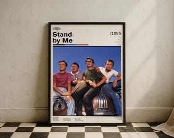 May include: A framed poster featuring the movie "Stand by Me" with the title and the year 1986. The poster shows four young boys sitting on a car with a blue sky background. The poster is in a black frame.