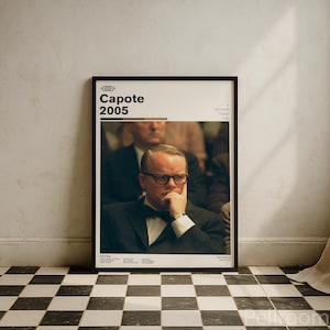May include: Framed movie poster for "Capote" (2005) featuring a portrait of Philip Seymour Hoffman. The poster has a black frame and is set against a white wall above a black and white checkered floor.
