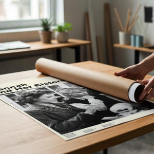 K&ouml;nnte beinhalten: Ein Schwarz-Wei&szlig;-Poster mit dem Titel "Hannah and Her Sisters" wird aus einer braunen Papprolle gerollt. Das Poster zeigt eine Vintage-Fotografie von zwei Frauen und einem Sch&auml;del. Das Poster liegt auf einem Holztisch.