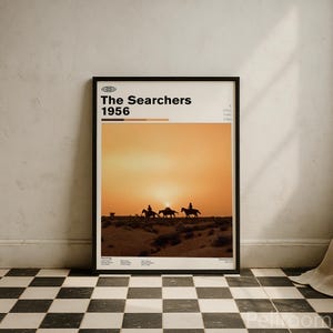 May include: Framed poster featuring the movie title "The Searchers 1956" with a sunset scene and silhouettes of riders on horseback. The poster has a black frame and is displayed against a white wall above a black and white checkered floor.
