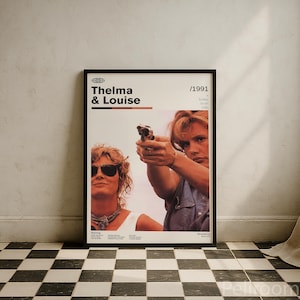 Puede incluir: Póster enmarcado de la película "Thelma & Louise" con el título en letras blancas en negrita. El póster presenta a dos mujeres, una de ellas apuntando con un arma. El póster está en un marco negro y se muestra contra una pared blanca.