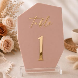 May include: A dusty rose acrylic table number sign with an irregular geometric shape. The sign features the word Table in gold cursive script above the large gold numeral 1. The sign is supported by a clear acrylic stand on a flat surface.