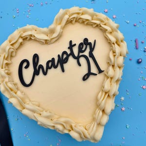 May include: A heart-shaped cake with a cream-coloured frosting base and a decorative border of piped buttercream. The cake features the words "Chapter 21" in black script. Sprinkles and a blue background complete the celebratory design.