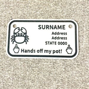 May include: A white rectangular sign with rounded corners, featuring a crab illustration and the text "SURNAME", "Address", "STATE 0000", and "Hands off my pot!" in black. The sign has two small holes at the top.