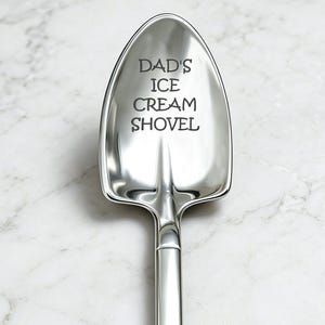 May include: A shiny, silver ice cream shovel with the text "DAD'S ICE CREAM SHOVEL" engraved on the shovel head. The shovel has a long handle and a triangular opening at the end. The background is a white marble surface.