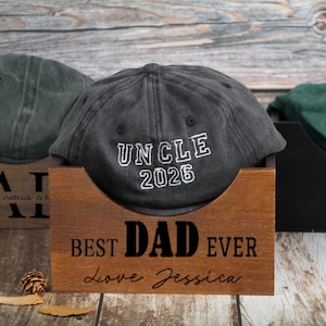 Puede incluir: Tres gorras de béisbol en cajas de madera. La gorra central es de color gris oscuro con "UNCLE 2026" bordado en blanco. La caja de abajo dice "BEST DAD EVER Love Jessica". Otras gorras son verdes y grises.