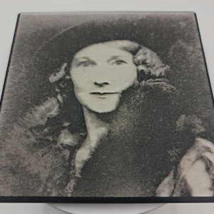 May include: A square, monochrome image of a woman in a hat and dark coat. The portrait has a vintage aesthetic, with a soft focus and a slightly grainy texture. The woman has long hair and a serious expression.