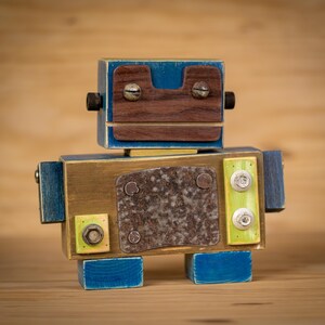 May include: A wooden robot toy painted in blue, brown and yellow. The robot has a face with two screws for eyes and a metal plate for a mouth. The robot has a rusty metal plate on its chest.