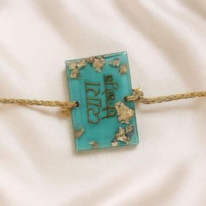 Puede incluir: Un colgante rectangular turquesa con detalles de pan de oro y texto dorado. El colgante está sujeto a un cordón trenzado de color dorado. El texto del colgante dice "Pyara Bhaiya".