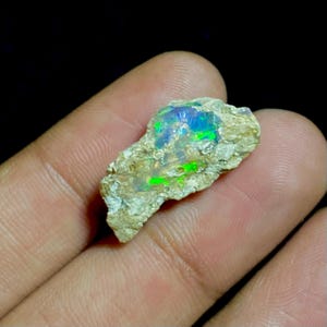 以下が含まれることがあります： 鮮やかな緑と青の輝きを持つ、未加工のオパール原石。 オパールはニュートラルな色調のマトリックスにセットされ、イリデッセントな遊色効果を際立たせています。 石は手で持たれており、そのサイズと自然な形を示しています。
