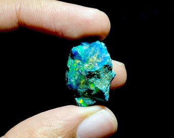 Puede incluir: Una piedra preciosa de ópalo en bruto, sin cortar, con vibrantes destellos azules, verdes y amarillos. El ópalo se sostiene entre dos dedos sobre un fondo negro, mostrando su forma natural e irregular y su juego de colores iridiscentes.