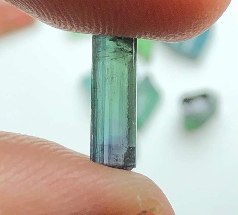 May include: A close-up of a tourmaline crystal held between two fingers. The crystal is a vibrant blend of green and blue hues, with a cylindrical shape and a slightly rough texture. Other small, similar crystals are visible in the background.