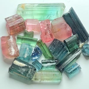 May include: A collection of raw tourmaline crystals in various colors, including pink, green, blue, and teal. The crystals are of different sizes and shapes, with visible natural textures and striations. The gemstones are arranged on a white surface.
