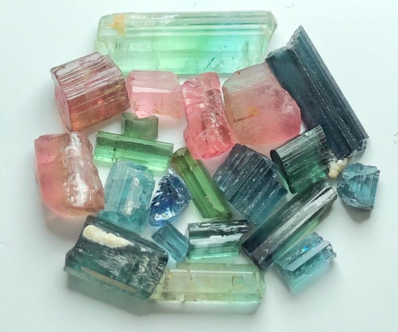 May include: A collection of raw tourmaline crystals in various colors, including pink, green, blue, and teal. The crystals are of different sizes and shapes, with visible natural textures and striations. The gemstones are arranged on a white surface.