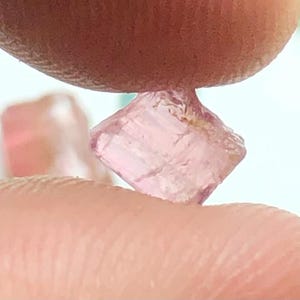 May include: A small, light pink, raw gemstone held between two fingers. The gemstone has a crystalline structure and a slightly translucent appearance. The background is a soft white, with hints of other gemstones.