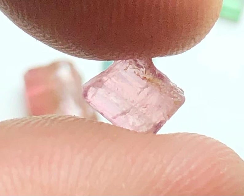 May include: A small, light pink, raw gemstone held between two fingers. The gemstone has a crystalline structure and a slightly translucent appearance. The background is a soft white, with hints of other gemstones.