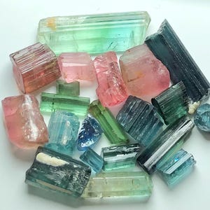 May include: A collection of raw tourmaline crystals in various colors, including pink, green, blue, and teal. The crystals are of different sizes and shapes, with a natural, rough texture. The arrangement is scattered, showcasing the gemstones' natural beauty.