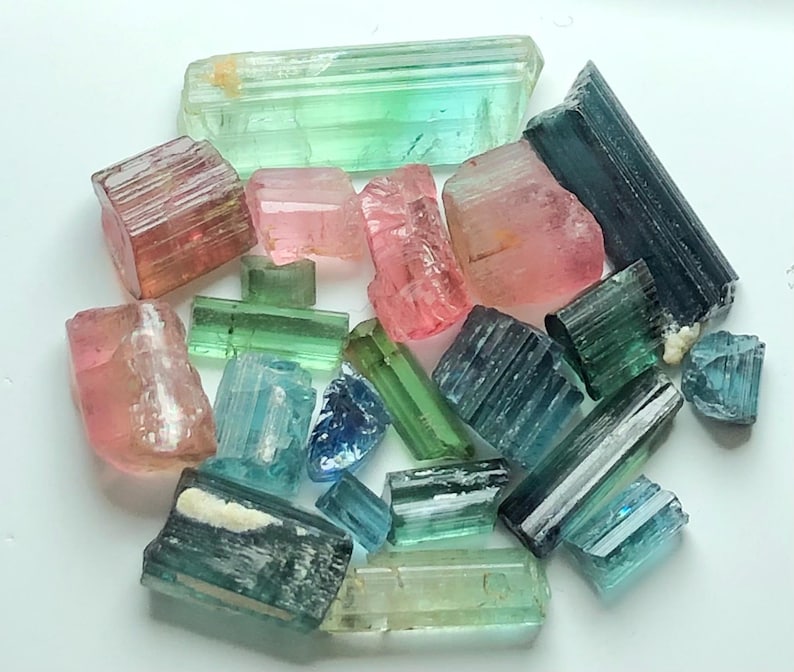 May include: A collection of raw tourmaline crystals in various colors, including pink, green, blue, and teal. The crystals are of different sizes and shapes, with a natural, rough texture. The arrangement is scattered, showcasing the gemstones' natural beauty.