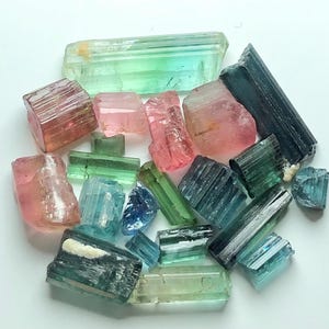 May include: A collection of raw tourmaline crystals in various colors, including pink, green, blue, and teal. The crystals are of different sizes and shapes, with visible natural textures and striations. The gemstones are scattered on a white surface.