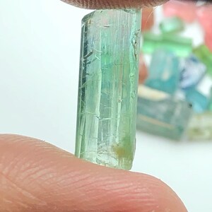 May include: A translucent, light green gemstone held between fingers. The crystal is a rectangular prism with visible internal striations. Other colorful gemstones are blurred in the background.