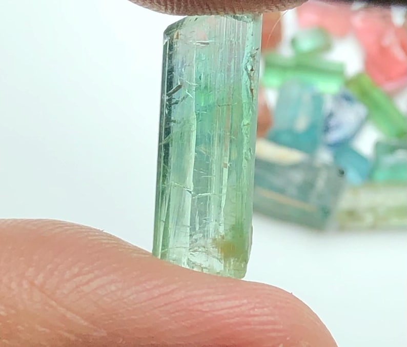 May include: A translucent, light green gemstone held between fingers. The crystal is a rectangular prism with visible internal striations. Other colorful gemstones are blurred in the background.