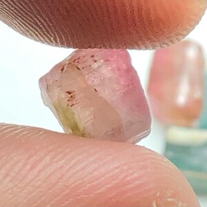 May include: A rough-cut, translucent gemstone with pink and yellow hues is held between two fingers. The gem has a crystalline structure and is slightly irregular in shape. Other gemstones are visible in the background.