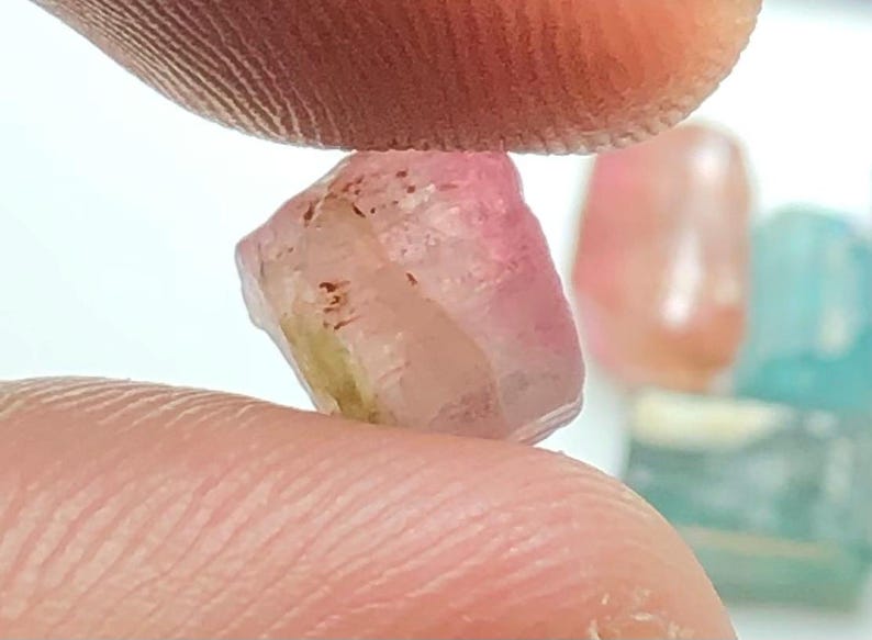 May include: A rough-cut, translucent gemstone with pink and yellow hues is held between two fingers. The gem has a crystalline structure and is slightly irregular in shape. Other gemstones are visible in the background.