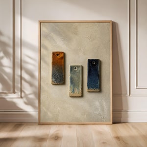 Op de afbeelding: Een ingelijst kunstwerk aan de muur met drie rechthoekige keramische tegels met elk een gat aan de bovenkant. De tegels zijn verticaal gerangschikt en tonen een verloop van gebrand oranje, lichtblauw en donkerblauw glazuur op een beige achtergrond.
