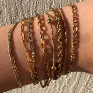 Pode incluir: Uma coleção de pulseiras douradas em vários estilos de corrente, incluindo designs de cobra, elos e corda. As pulseiras são exibidas em um pulso, mostrando as diferentes texturas e padrões de cada peça. As joias são provavelmente feitas de metal.