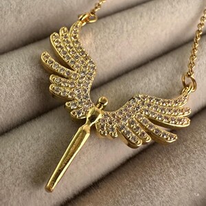 May include: A gold-colored necklace with an angel wing pendant. The pendant is adorned with small, clear gemstones. The necklace chain is also gold-colored. The necklace is displayed on a soft, gray surface.