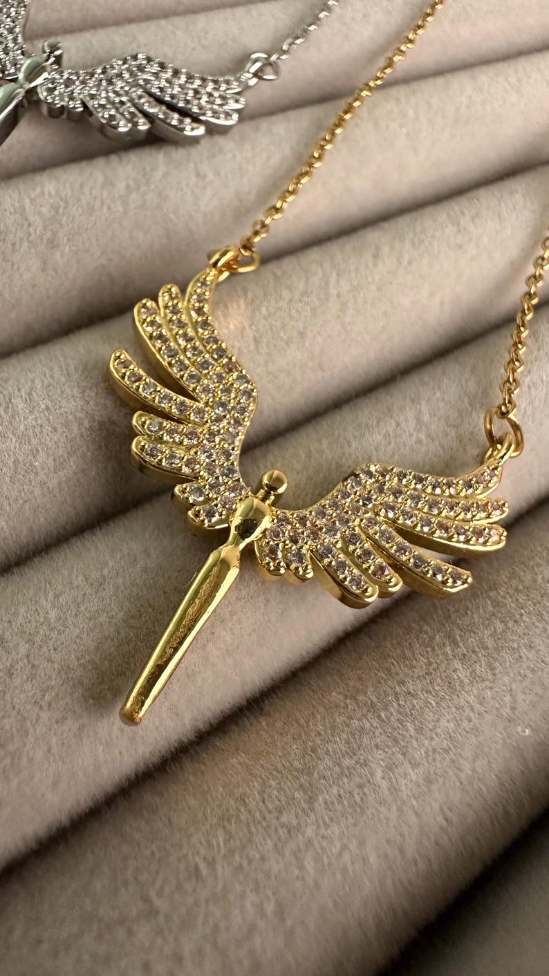 May include: A gold-colored necklace with an angel wing pendant. The pendant is adorned with small, clear gemstones. The necklace chain is also gold-colored. The necklace is displayed on a soft, gray surface.