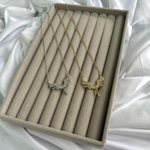 May include: Two necklaces with angel wing pendants. One necklace is silver-toned, and the other is gold-toned. Both pendants feature a detailed wing design with small, sparkling embellishments. The necklaces are displayed on a gray velvet surface.