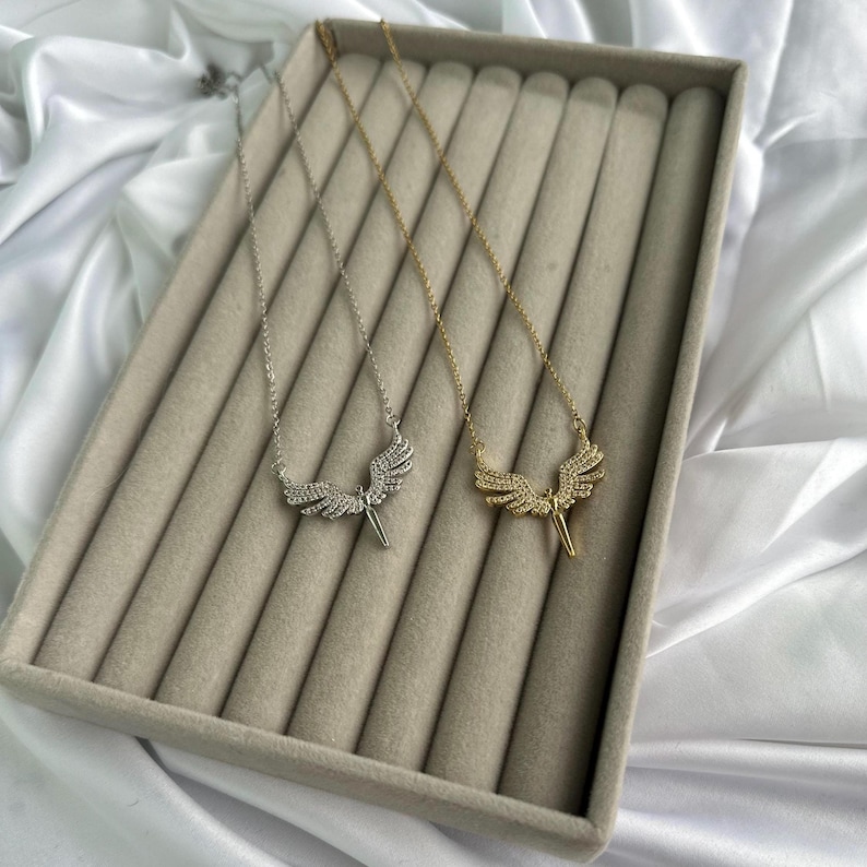 May include: Two necklaces with angel wing pendants. One necklace is silver-toned, and the other is gold-toned. Both pendants feature a detailed wing design with small, sparkling embellishments. The necklaces are displayed on a gray velvet surface.