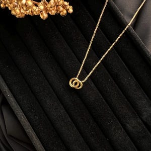 May include: A gold necklace featuring a fine chain and a pendant of two interlocking circles. The necklace is presented on a black velvet surface, with a gold floral decoration in the background.