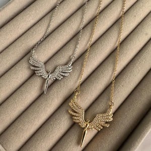 May include: Two necklaces with angel wing pendants. One necklace is silver-toned, the other is gold-toned. Both pendants feature a figure with outstretched wings, encrusted with small, sparkling stones. The necklaces rest on a beige, textured surface.