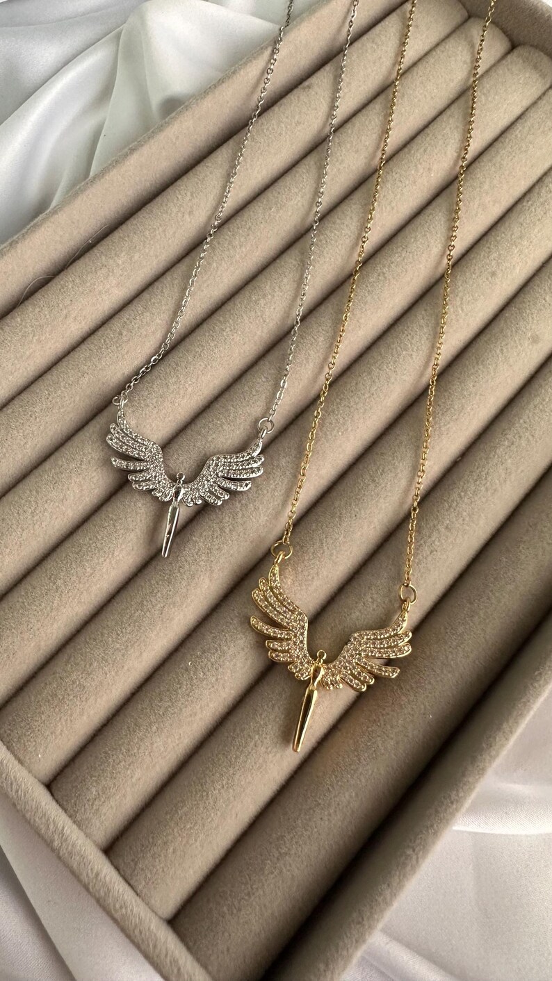May include: Two necklaces with angel wing pendants. One necklace is silver-toned, the other is gold-toned. Both pendants feature a figure with outstretched wings, encrusted with small, sparkling stones. The necklaces rest on a beige, textured surface.