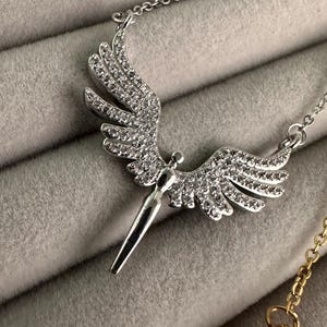 May include: Silver-toned necklace featuring a pendant shaped like angel wings. The wings are encrusted with small, sparkling crystals. The pendant has a stylized figure at the center and hangs from a delicate chain. The background is a soft, gray fabric.