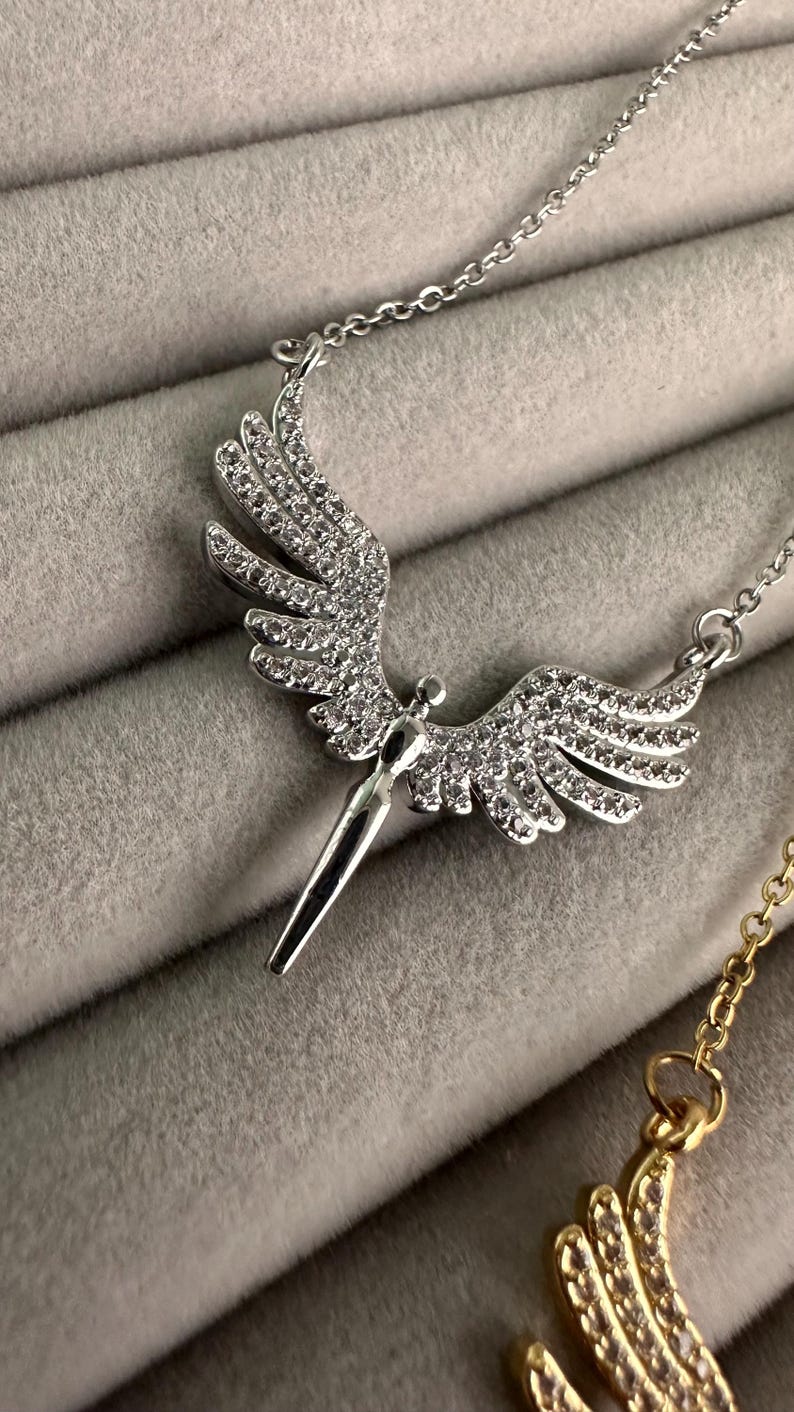 May include: Silver-toned necklace featuring a pendant shaped like angel wings. The wings are encrusted with small, sparkling crystals. The pendant has a stylized figure at the center and hangs from a delicate chain. The background is a soft, gray fabric.