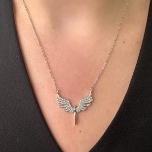 May include: A silver-toned necklace featuring a pendant shaped like angel wings. The wings are embellished with small, sparkling stones. The necklace is displayed against a dark background, highlighting the jewelry's details.