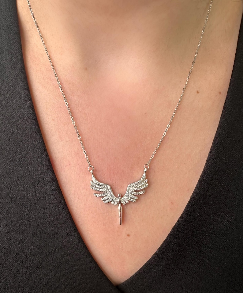 May include: A silver-toned necklace featuring a pendant shaped like angel wings. The wings are embellished with small, sparkling stones. The necklace is displayed against a dark background, highlighting the jewelry's details.