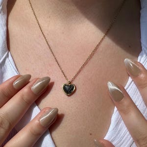 May include: A gold-colored necklace with a heart-shaped pendant. The pendant has a dark, possibly black, center and a gold-colored border. The necklace is on a person wearing a white shirt. The person's hands with long, polished nails are visible.