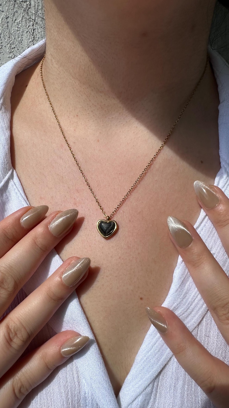 May include: A gold-colored necklace with a heart-shaped pendant. The pendant has a dark, possibly black, center and a gold-colored border. The necklace is on a person wearing a white shirt. The person's hands with long, polished nails are visible.