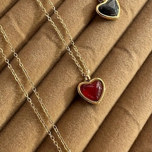 May include: Gold-toned necklaces with heart-shaped pendants. One pendant is black, one is red, and one is white. Each heart is encased in a gold frame and hangs from a delicate chain. The necklaces are displayed on a brown, textured surface.