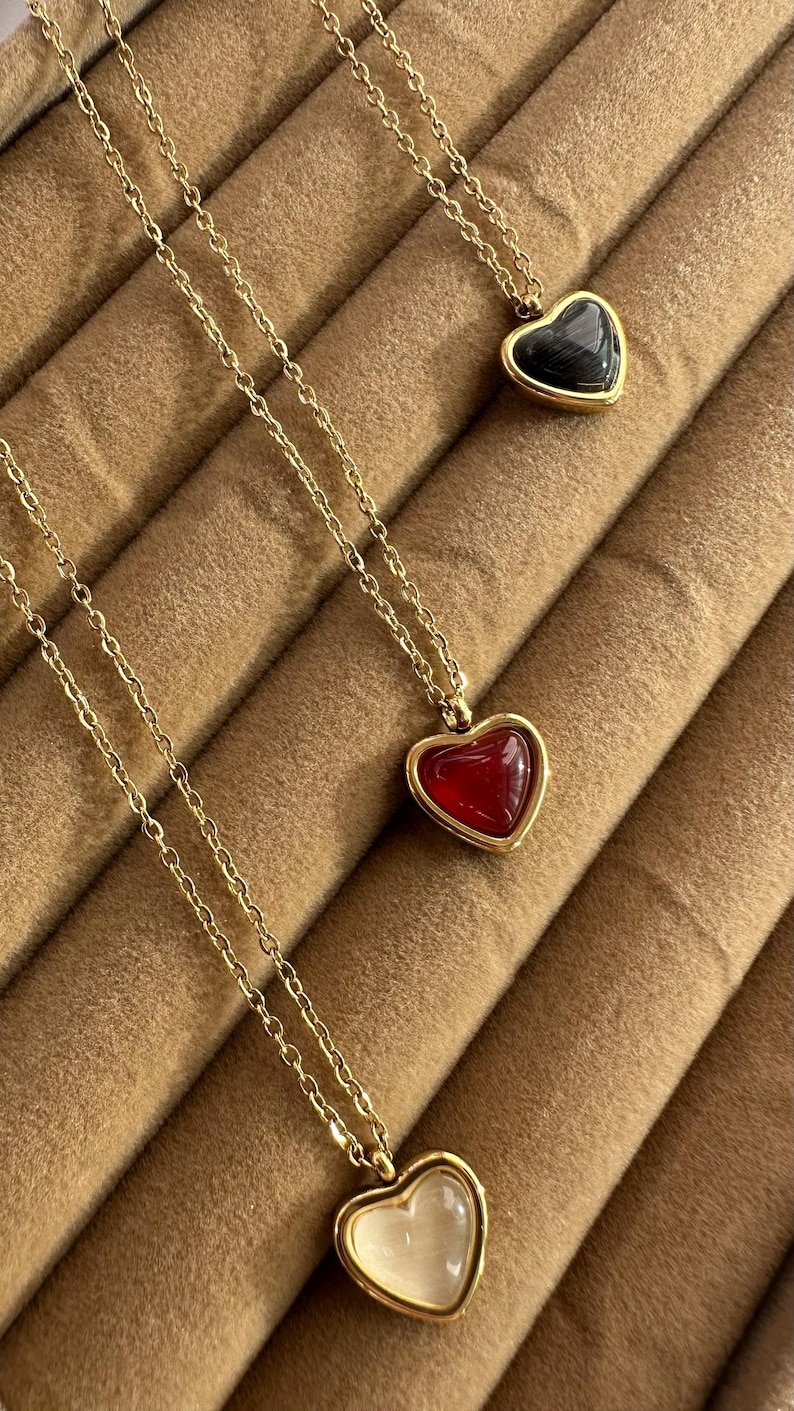 May include: Gold-toned necklaces with heart-shaped pendants. One pendant is black, one is red, and one is white. Each heart is encased in a gold frame and hangs from a delicate chain. The necklaces are displayed on a brown, textured surface.
