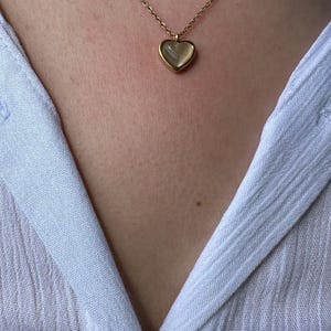 May include: A gold-colored necklace with a heart-shaped pendant. The pendant has a light-colored, possibly mother-of-pearl, center. The necklace is worn against a light blue shirt.